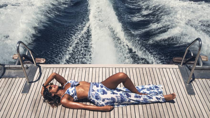 Woman sunbathes on the deck of a boat, the frothy and foaming wake visible for miles