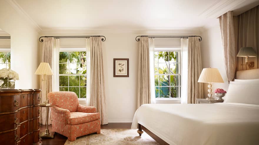 Wood dresser, pink floral armchair by tall sunny windows, nightstand and bed