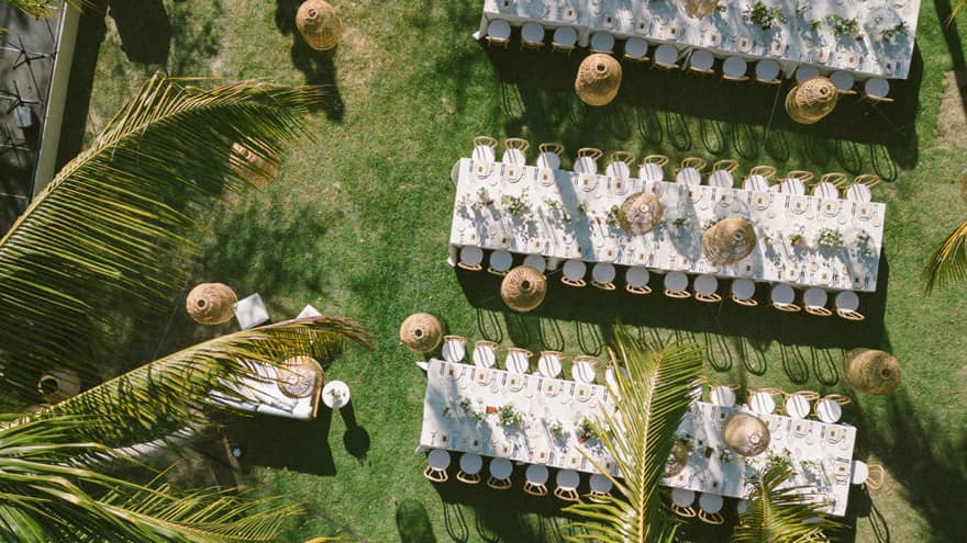An aerial view of an outdoor event setup on a green lawn surrounded by palm trees. Setup includes long dining tables set for an event, chairs, lighting and other decor.