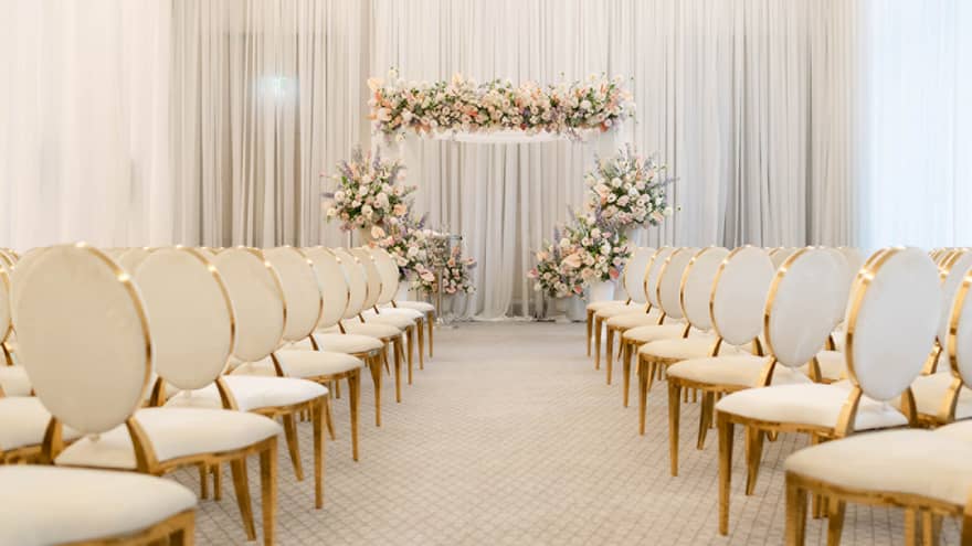 A stunningly decorated wedding aisle flanked by beautifully arranged cream and gold chairs, leading up to flower arch at altar, ready to welcome guests