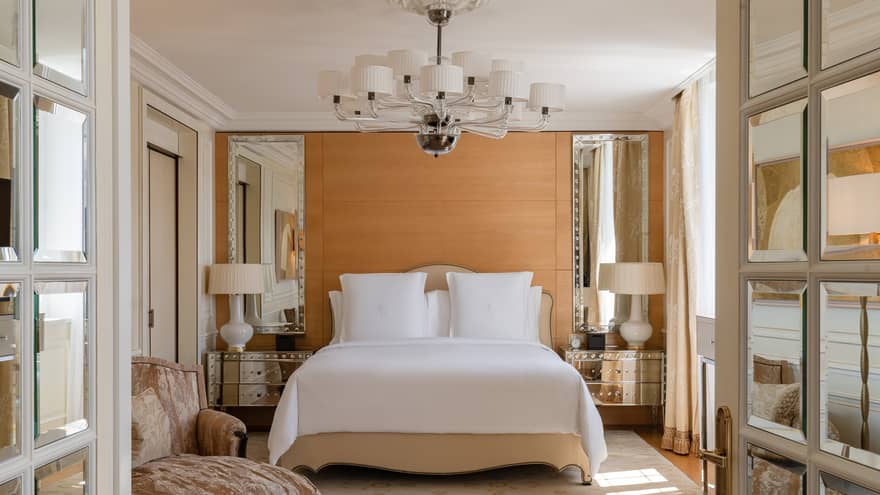 Luxury hotel bedroom with a king bed, mirrored bedside tables with lamps and a modern chandelier, complemented by soft beige accents and an elegant armchair