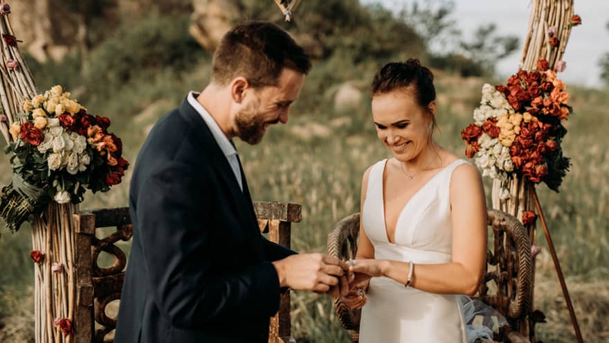 Groom smiling, placing ring on smiling bride's finger in archway on Serengeti bushland