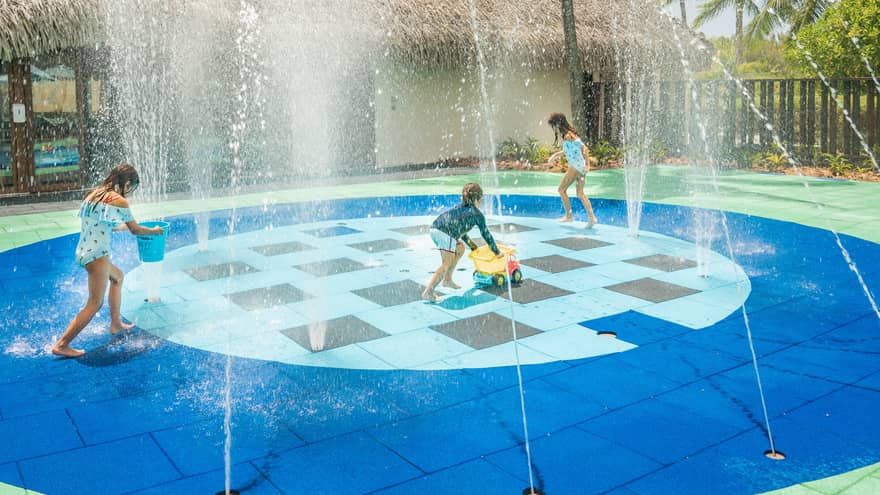 Group of children in swimsuits play on splash pad next to grass-roof building