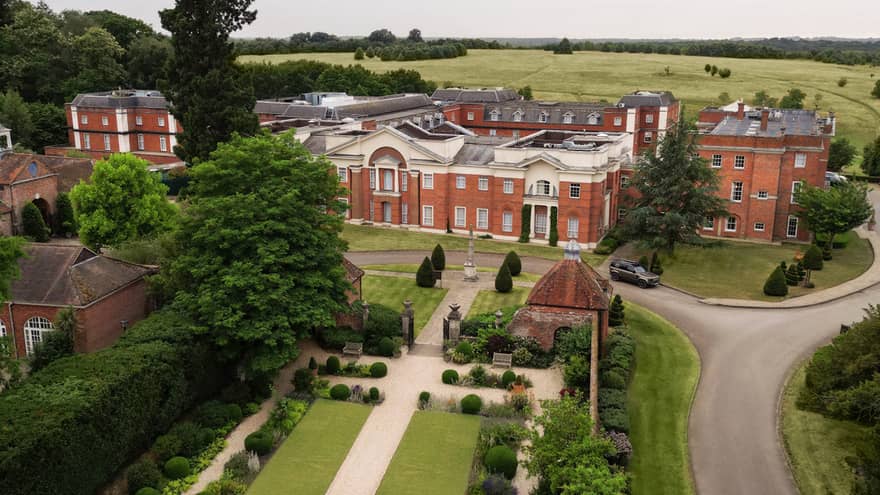 A bird's-eye view of a property with a large structure, a manicured garden, wooded areas and fields in the background
