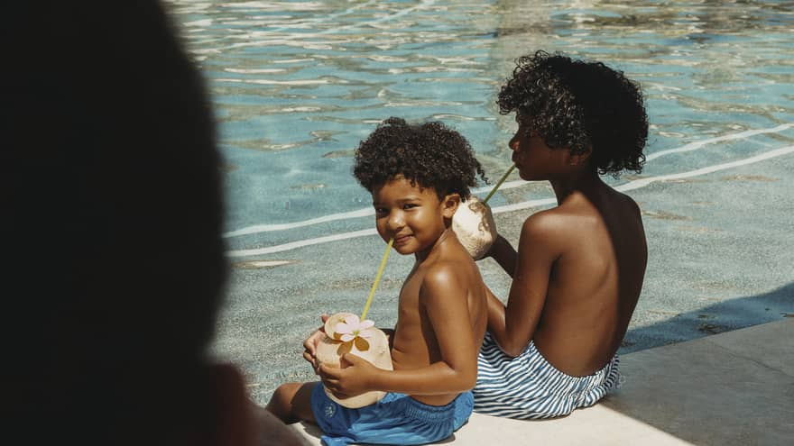 Two children sitting by a poolside drinking from coconuts with straws on a sunny day