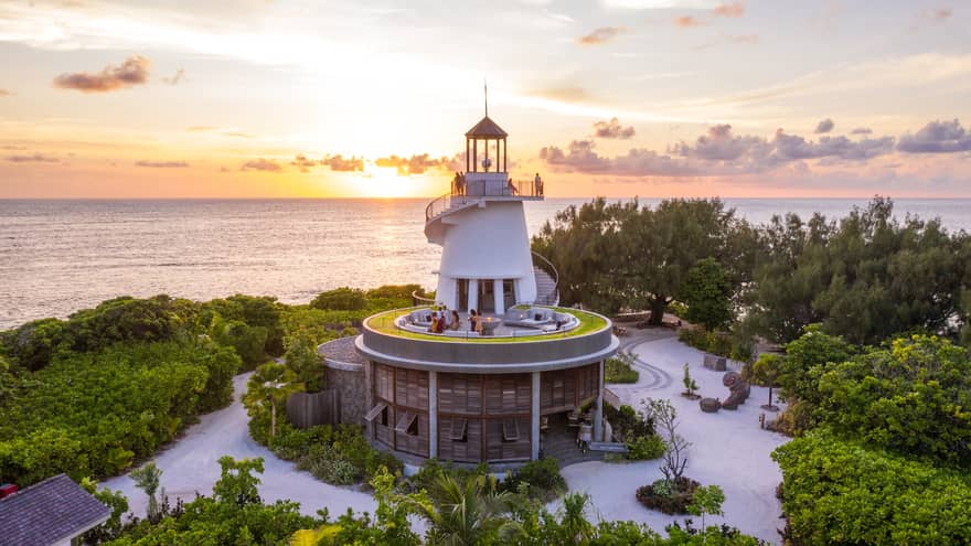 Lighthouse nestled among greenery with people on different levels, overlooking the ocean at sunset