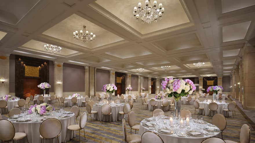 A chandelier-clad ballroom set with several circular tables boasting tall displays of hydrangeas. 