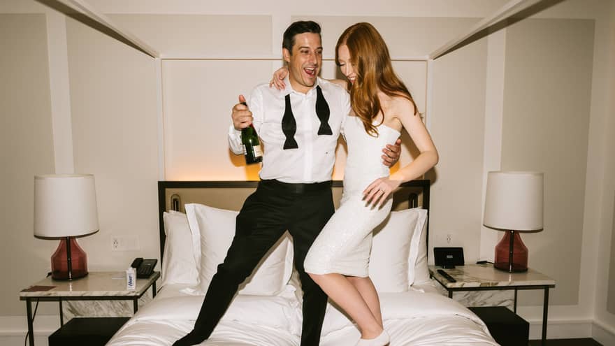 A stylish couple, dressed in an elegant white dress, shirt and black pants, joyfully stand on a bed, celebrating with a bottle of champagne