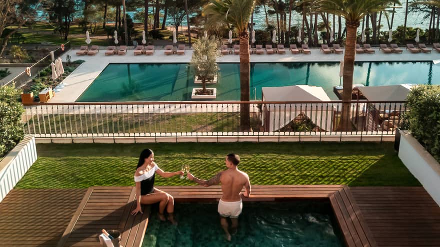 Two people in a small square pool in an outdoor setting. A larger pool surrounded by lounge chairs is in the background, as are ocean and mountain views beyond that. Verdant lawns and trees surround the pool areas.