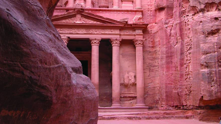 Light filters down on a sandstone tomb cut into a rock face hidden behind a large rock formation, highlighting its red tones.