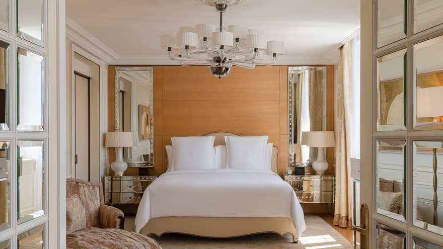 Luxury hotel bedroom with a king bed, mirrored bedside tables with lamps and a modern chandelier, complemented by soft beige accents and an elegant armchair
