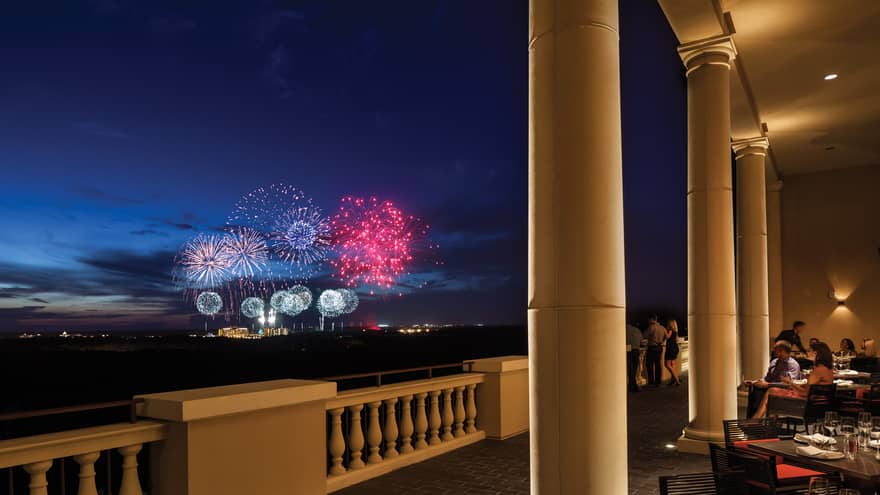 The terrace of a restaurant with tables and people watching fireworks in the distance.