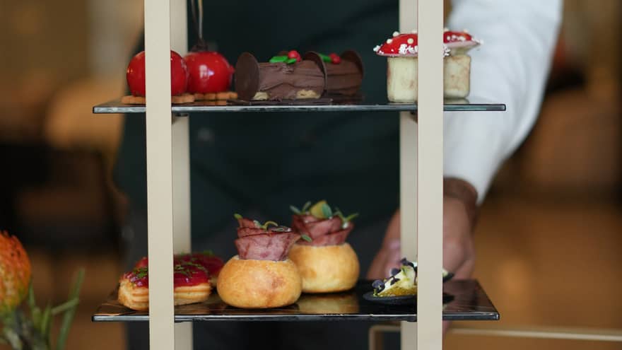 Server wearing black pants, black vest and white button-down shirt presents a three-tiered tray of afternoon tea desserts
