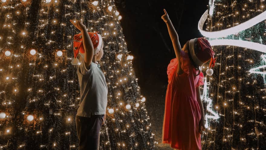 Two children wearing Santa hats, standing in front of two lit Christmas trees with one hand pointed upwards