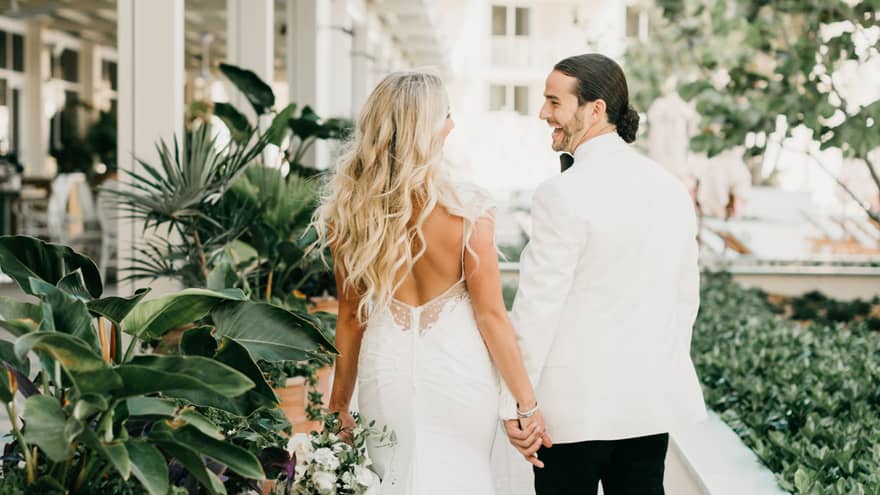 A bride and groom strolls on a lush outdoor patio, surrounded by vibrant plants in clay pots.