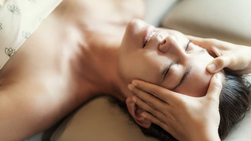 Spa facial, two hands rest on woman's forehead as she closes her eyes, lays under sheet on treatment table