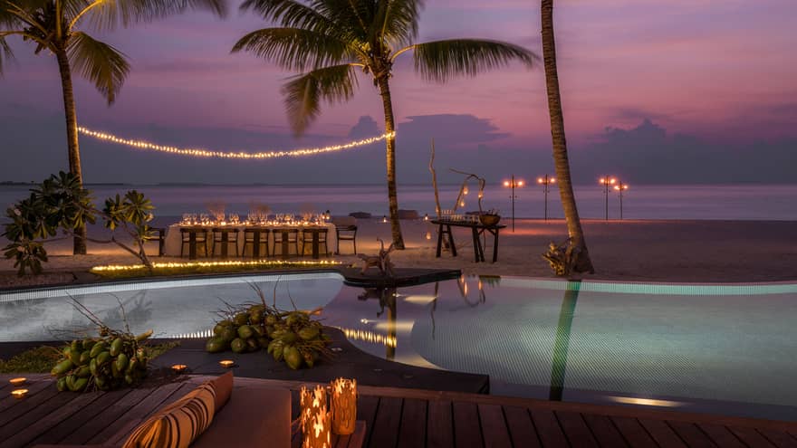 String of lights between palm tree over private dining table on beach by swimming pool at sunset