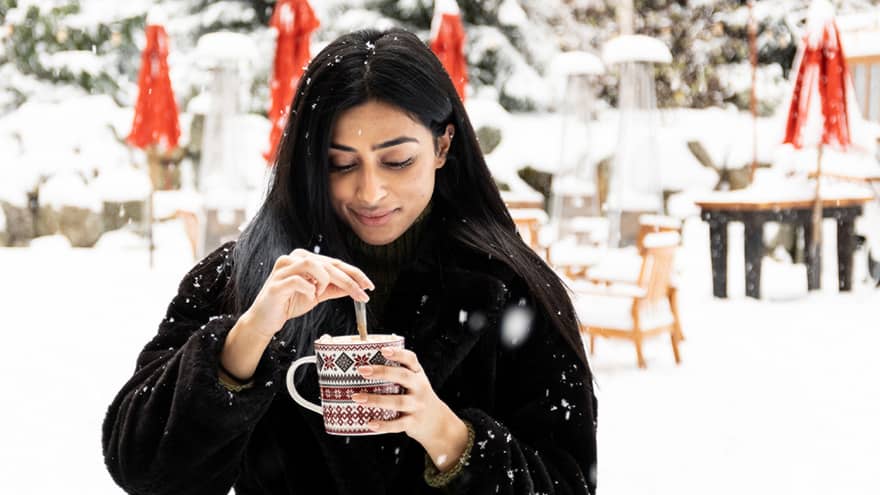Woman in black coat stirring drink in a festive mug with snow-covered trees and tables in backdrop