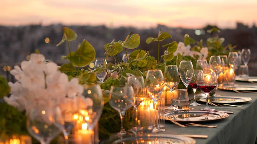 Outdoor table with teal linen, white-and-gold plates, gold cutlery, white-and-green floral centrepieces and lit candles all set beneath a pink-and-=white sky as the sun sets over the city