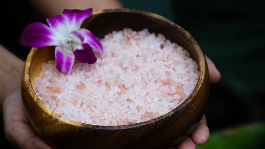 Two hands hold a wooden bowl filled with pink salt and a purple flower