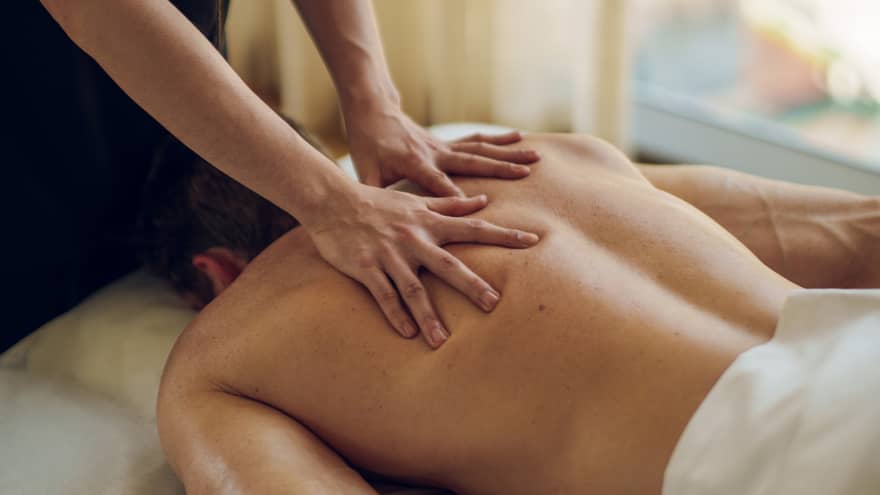 Guest lays face down on a massage table covered in a blanket from the waist down while a massage therapist massages his back