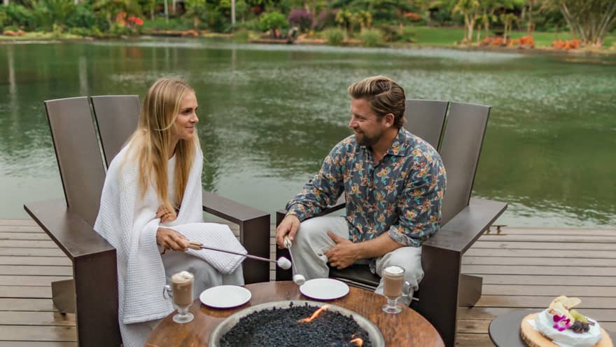 Man and woman roast marshmallows on wooden deck by pond