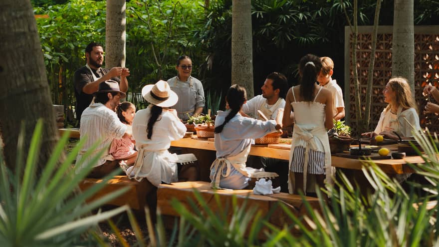 People seated around a long outdoor dining table set among palm trees