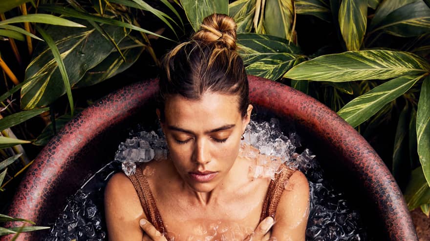 A guest, wearing a bathing suit, is sitting and relaxing in a rustic bathtub full of ice with foliage encircling the tub.