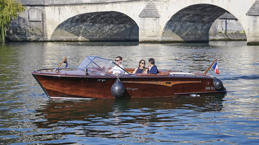 A couple relax in a brown speedboat cruising near a stone bridge along a river lined with white Haussmannian-style buildings.