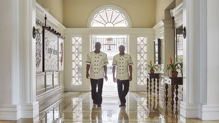 Two hotel staff wearing white uniforms walk across marble floors in sunny lobby
