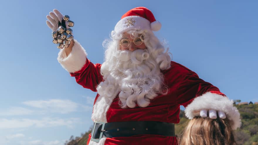 Person dressed in a red Santa Claus suit and white beard holds jingle bells in their hand and waves while standin next to a child on a beach