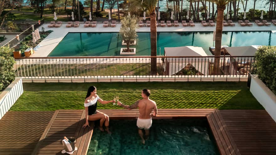 Two people in a small square pool in an outdoor setting. A larger pool surrounded by lounge chairs is in the background, as are ocean and mountain views beyond that. Verdant lawns and trees surround the pool areas.