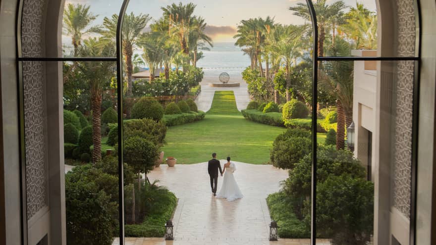 View through window looking out onto a bride and groom walk along a tiled outdoor pathway toward a green lawn with palm trees and the beach in the distance
