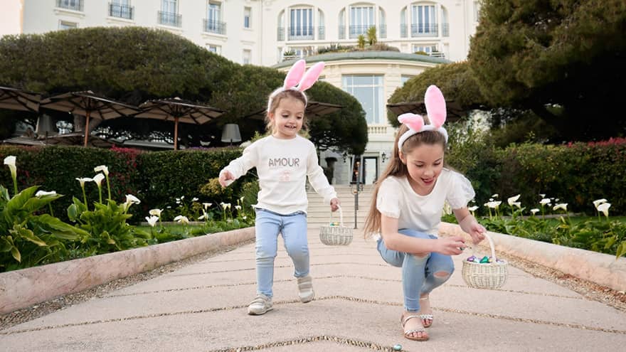 Two children in matching outfits wearing pink rabbit ears and holding Easter baskets with grand white building in backdrop