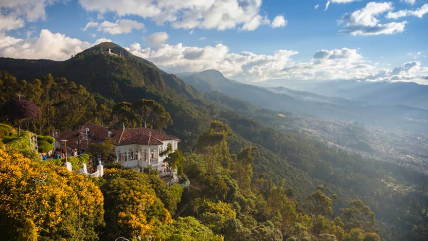 Aerial view of colonial-style white building tucked high on lush green mountain that stretches to horizon, blue sky