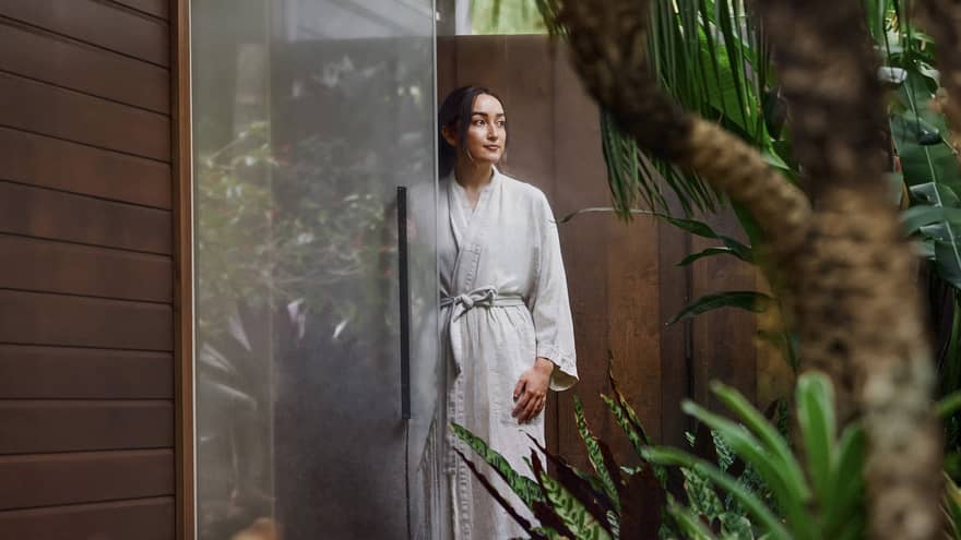 A woman dressed in a bathrobe stands by a glass door at an outdoor spa area surrounded by lush greenery