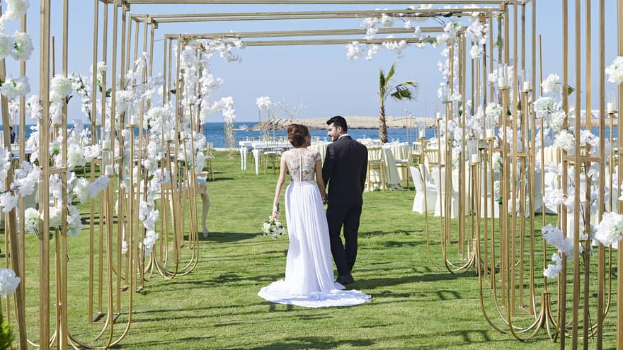 Rear view of wedding couple holding hands and smiling under a floral and gold archway path on lawn