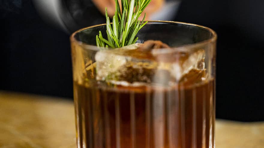 A dark amber coloured cocktail with an herb garnish.