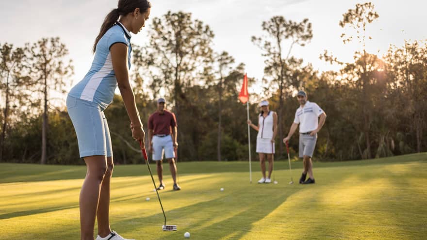 Woman with gold club prepares to putt as friends hold flag on golf course green