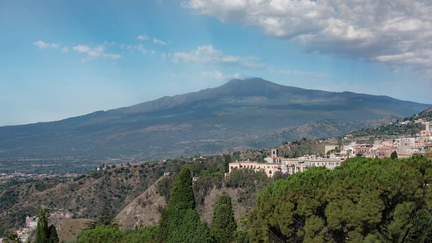 Hillside buildings surrounded by trees, Mount Etna and blue sky in backdrop