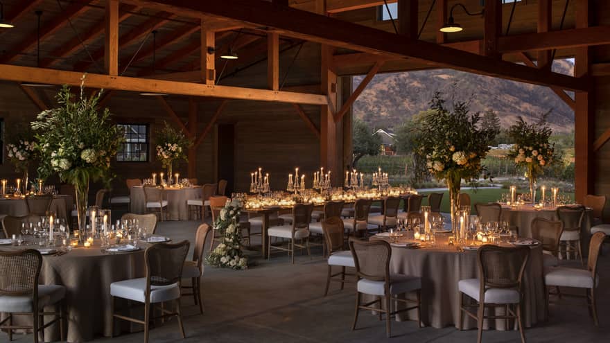 A beautifully arranged barn wedding reception, featuring tables and chairs under soft, dim lighting.