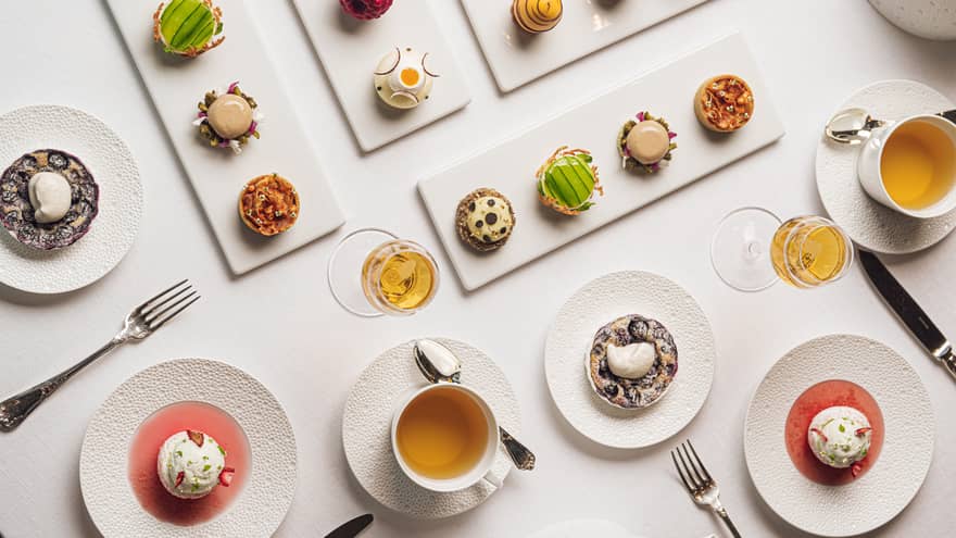 Four white rectangular platters each holding four colourful pastries each are set on a white table along with two cups of tea and four small round plates of dessert