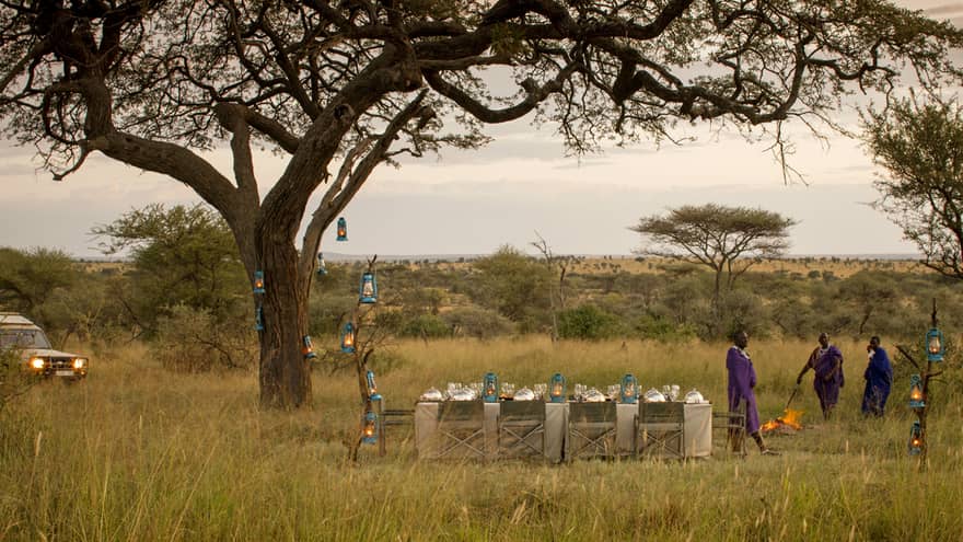 Three Maasai stand beside dining table and chairs, lanterns beside tree and jeep in Serengeti 