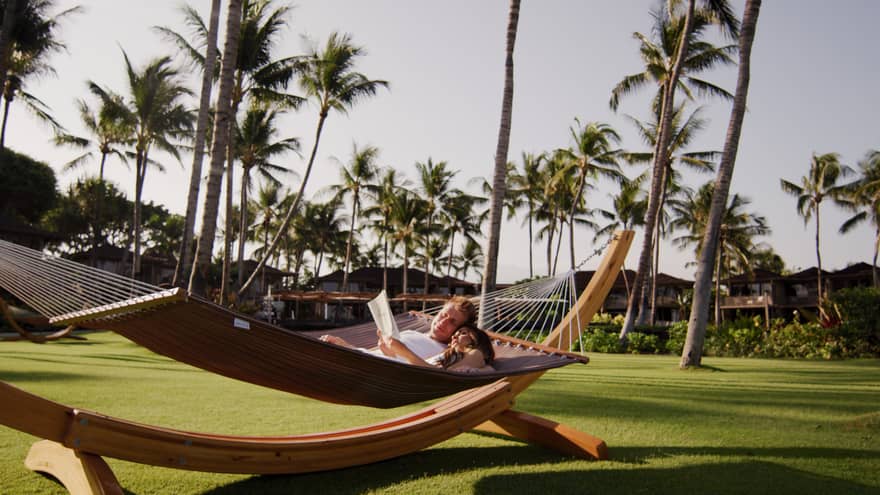 Two guesting laying down and reading in an outdoor hammock, surrounded by palm trees