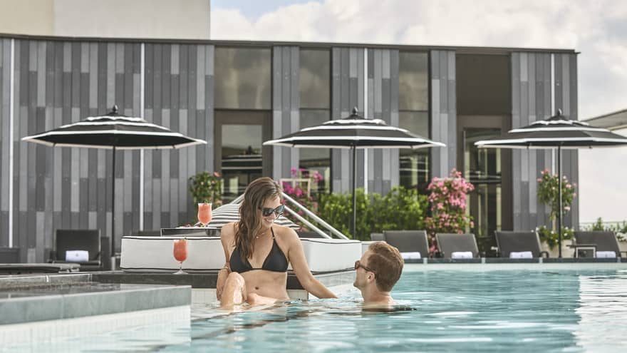 Relaxed couple enjoying a sunny day in the rooftop infinity pool at Four Seasons New Orleans, surrounded by modern lounge areas and elegant umbrellas, with a refreshing cocktail in view.