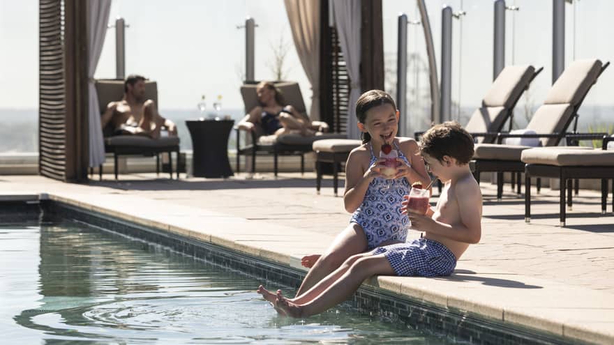 Two children sitting by the edge of a pool, enjoying drinks, while adults relax on lounge chairs under shaded cabanas in the background