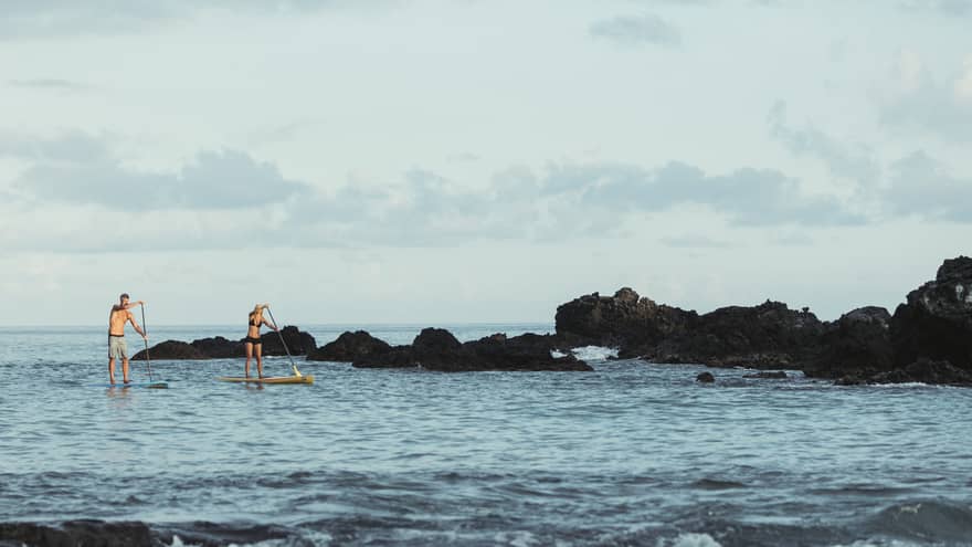 Two people balance on stand-up paddleboards in lagoon by black rocks