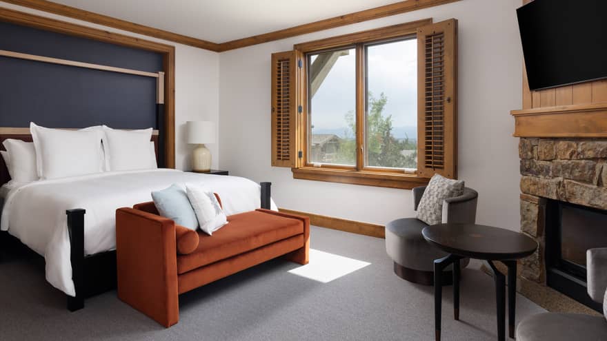 Luxury resort bedroom with a king bed, burnt orange chaise, grey armchairs, stone fireplace, TV and window with wooden shutters