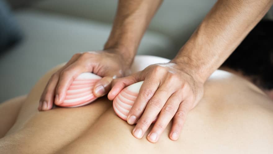 A masseuse's hands hold two white, pink-striped lava shells, running over the bare back of a man lying face down on a massage table