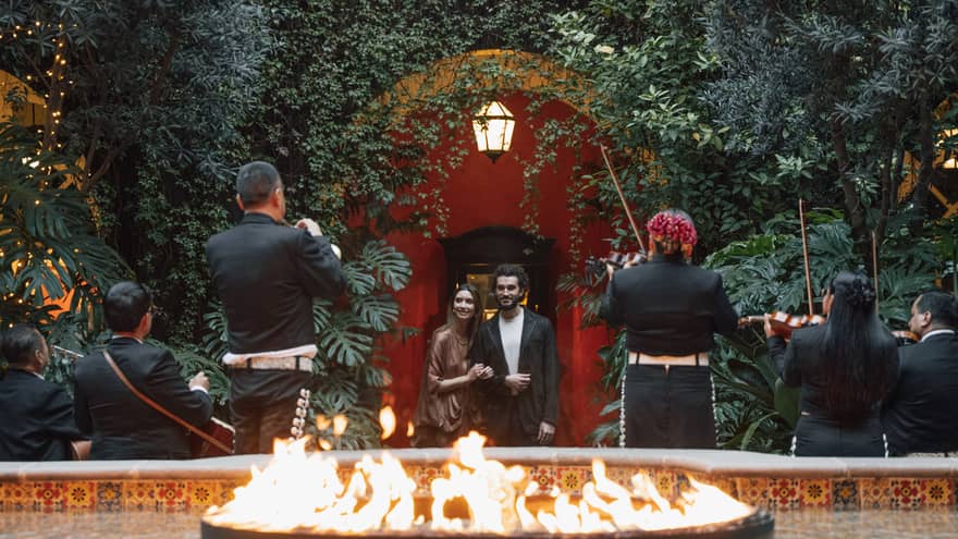 Two guests watch musicians play in a greenery-filled courtyard, with a fountain with a flame feature in the foreground.